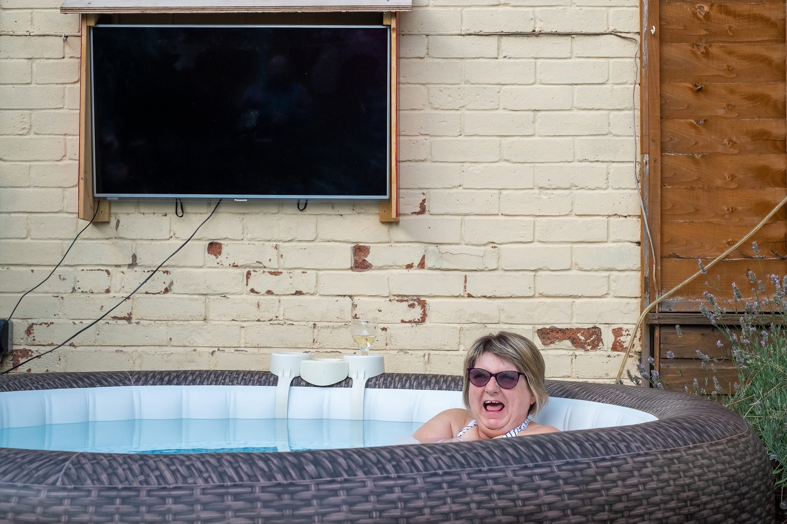 A white woman in a hot tub wearing sunglasses, only her head and shoulders in view, is laughing in the direction of the camera.