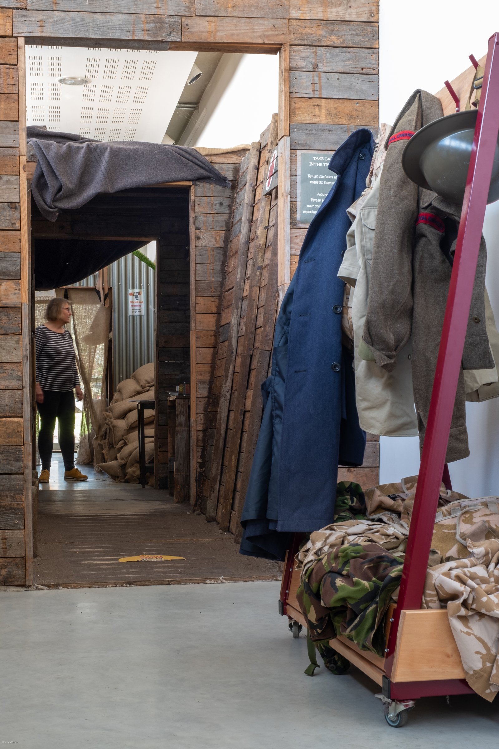 A white woman walks through a life-size air raid shelter and in the foreground is a display of military uniforms on a clothes rack.