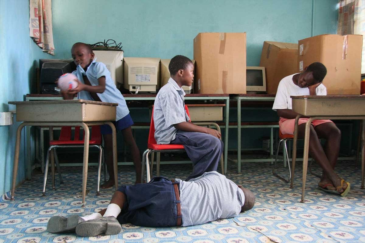 Four boys in school uniform in a classroom. One boy lies on the floor, another bounces a ball on the desk.