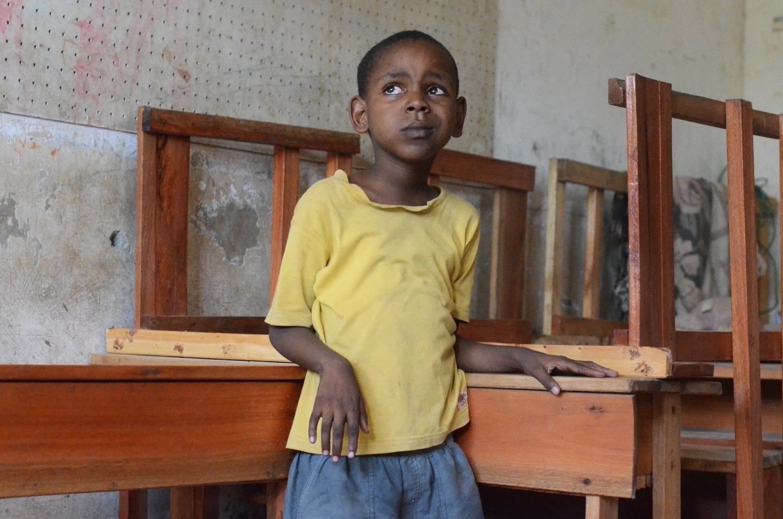 A boy in a yellow T-shirt glances upwards with an anxious expression. He leans on some school desks.