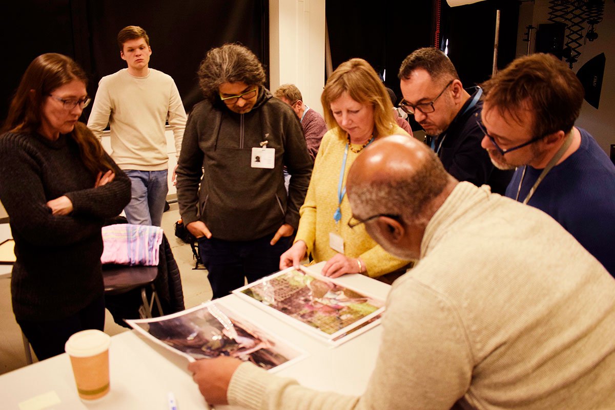People crowd around a table to look at A3-size photographs.