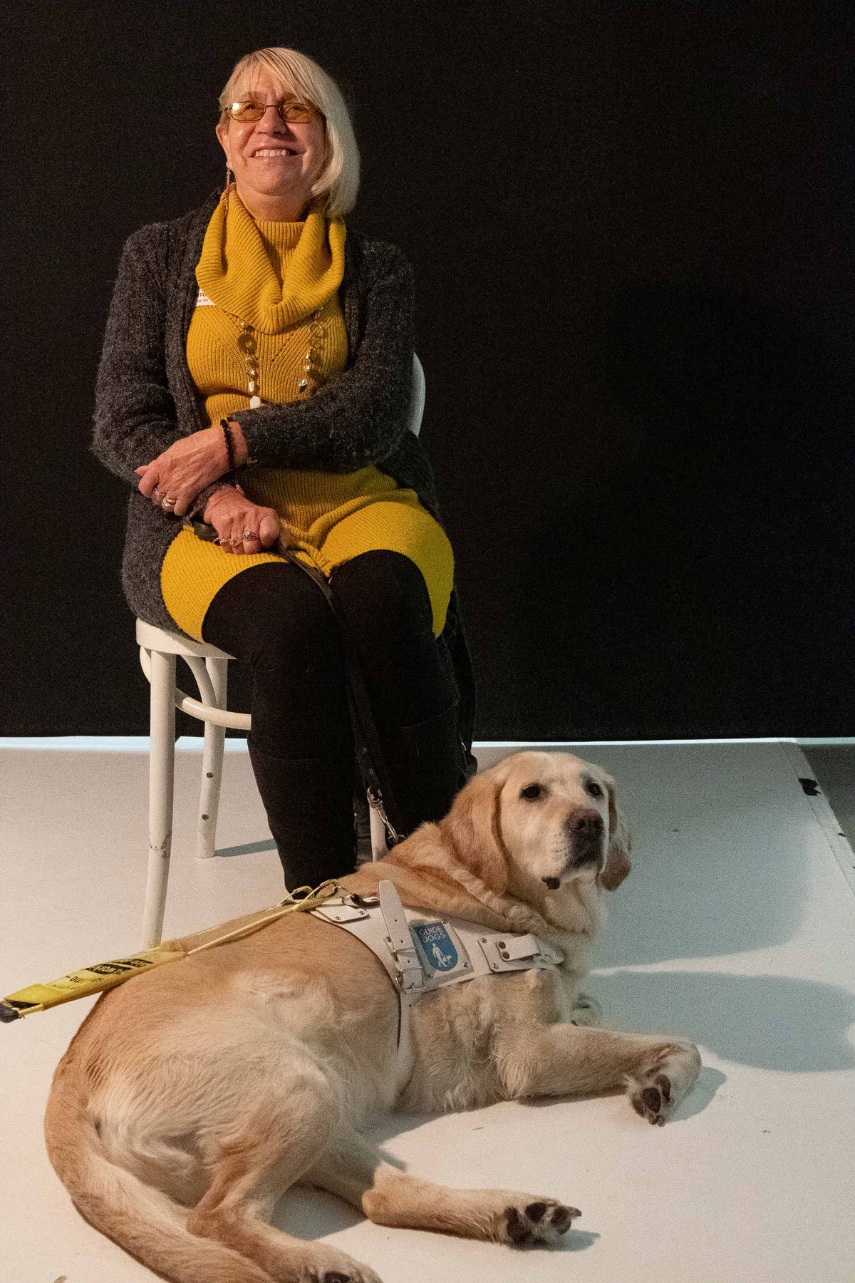 Studio photograph of a seated woman, smiling, with a blonde Labrador lying at her feet.