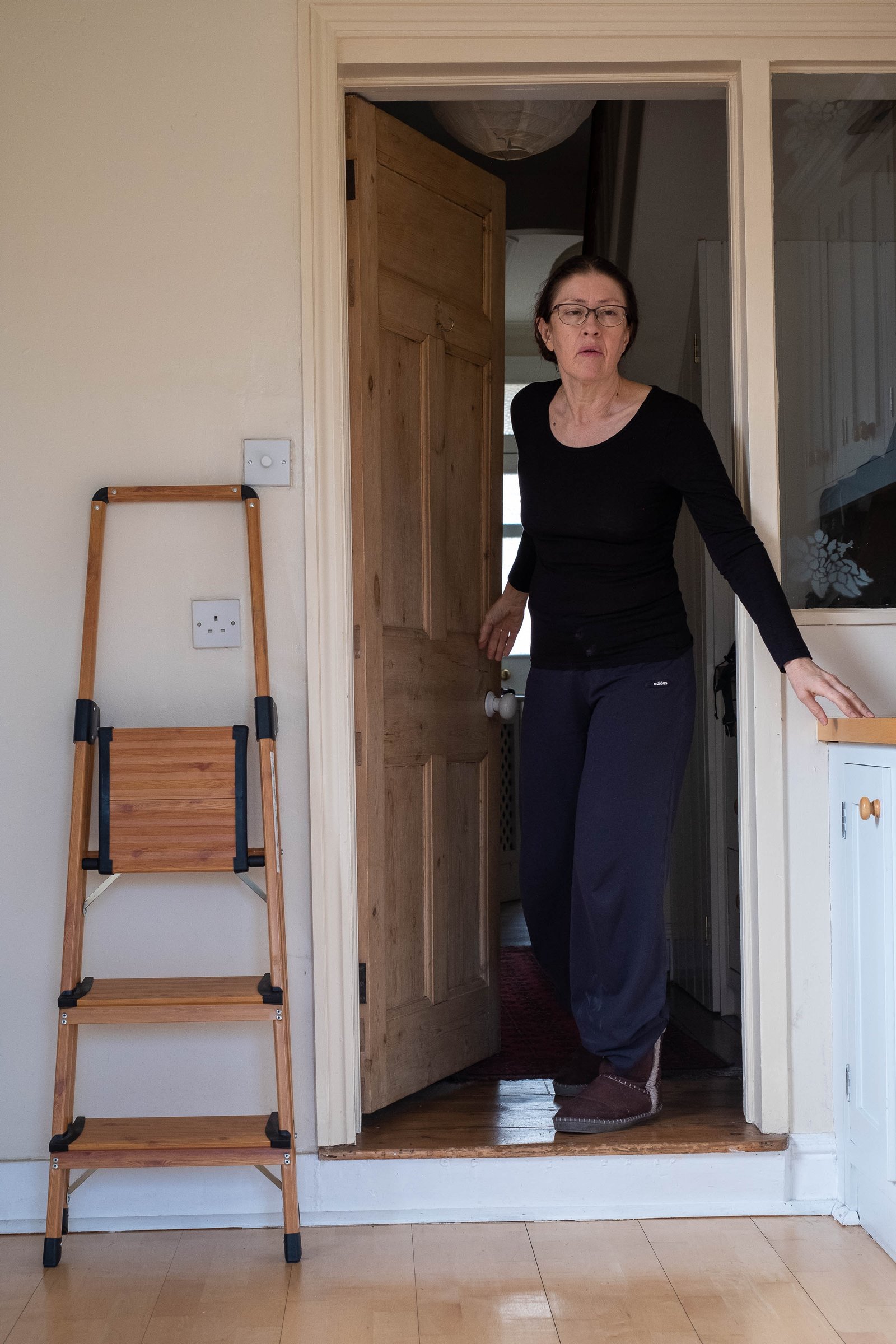 A woman in dark clothes steps through a door down into a kitchen. Her right hand touches a sideboard, a folded stepladder leans on the wall.