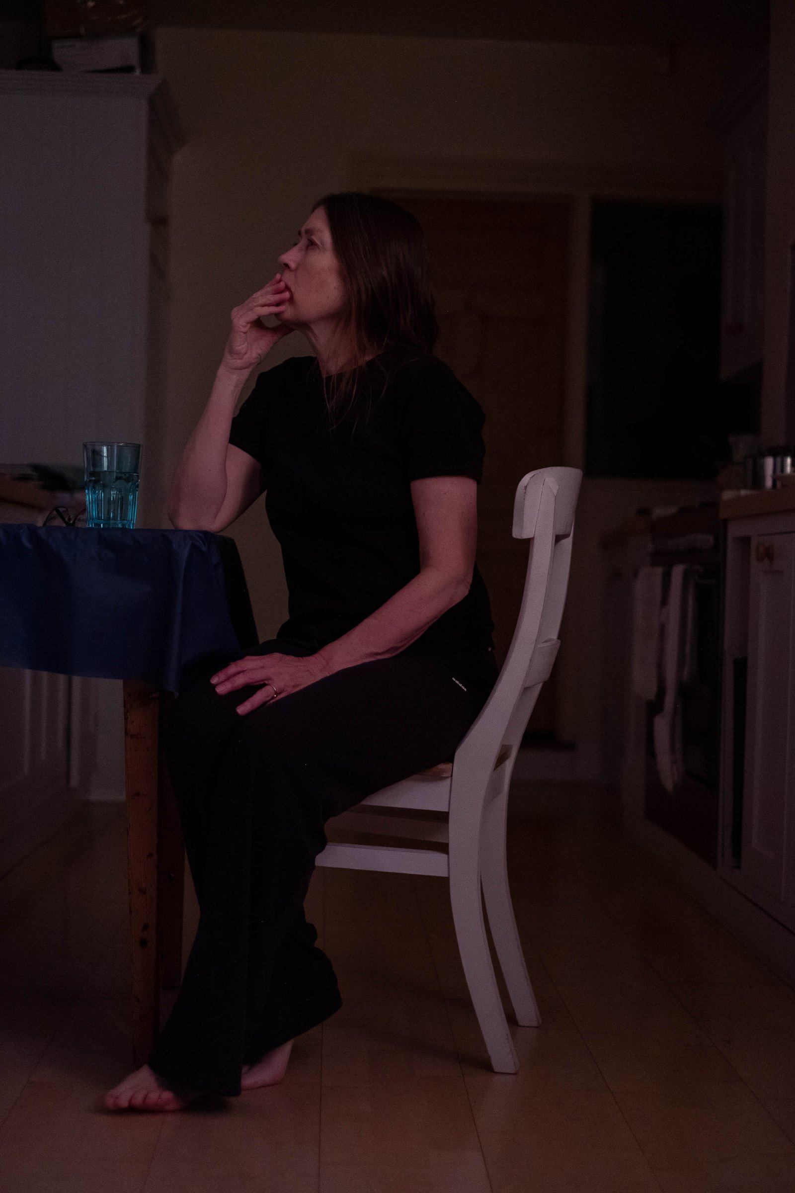 A woman dressed in black sits at a kitchen table in a dark room, gazing upwards as if lost in thought.